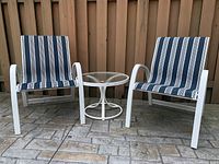 Two white metal patio chairs with blue and white striped fabric seats and backs, positioned on a stone patio next to a glass top circular white metal table. Visible rust on table base.