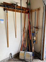 Full view of various yard tools including rakes, shovels, pitchfork, pickaxe, mop, dustpan all hanging on or leaning against garage wall
