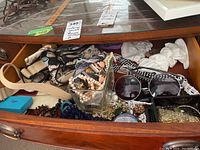 Open drawer showing various costume jewelry including beaded necklaces, bracelets, faux pearls in glass dish, fabric belt, and white hair berets