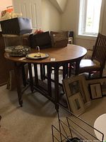 Round wooden gate leg dining table with one leaf attached, surrounded by one wooden chair with vinyl seat, in room corner with window and carpet.