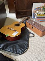 Acoustic guitar placed on or next to a black soft-sided case and wooden tongue drum beside it.