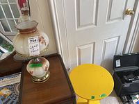 Two items on a wooden furniture piece near a white door: the glass lamp with floral design and the yellow metal side table.