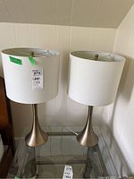 Pair of matching table lamps with white drum shades and brushed metal bases on a glass shelf, showing front view of both lamps.