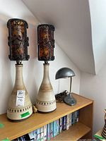 Full view of two matching ceramic table lamps with decorative amber-tinted glass shades held in black wrought iron frames, alongside one small gray metal adjustable desk lamp on a wooden shelf.