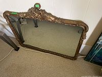 Framed wall mirror laid against a wall, showing full mirror and ornate gold frame with floral and scroll designs.