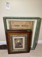 Three framed items leaning against a wall: one large framed newspaper article 'Golden Wedding', two smaller framed portraits with wooden and silver-toned trim frames