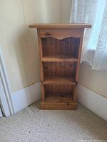 Wooden shelf unit positioned in corner near a window with lace curtains. Shows the front view of the empty shelf with wood grain and knots visible.