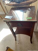 Front view of wood side table showing drawer, curved legs, and glass top reflecting window.