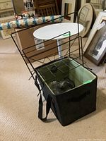 White round metal pedestal table with black metal frame magazine rack and black cloth bin with green lining placed nearby indoors on carpeted floor.