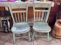 Pair of vintage wooden chairs with curved seats and spindle backs, painted in pale green with red stenciling, showing scratches and wear