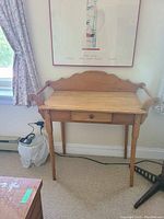 Front view of the wood wash stand against a wall, showing its rectangular top, curved arm supports, arched backboard, and single drawer below the tabletop.