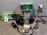 Wide view of lot showing Ricoh XR-7 camera with lens, multiple 35mm lenses, Akai microphones, aluminum tripod, and Pentax belt buckle on concrete floor.