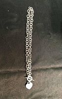 Full length of the silver chain necklace laid flat on a black background showing the toggle clasp and heart charm.
