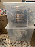 Two large clear plastic bins filled with assorted silver plate items including pitchers, serving vessels, and plates stacked inside at an angle.
