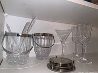 Two crystal ice buckets, one with cut pattern, one ribbed, both with metal rims and handles. Three crystal stem glasses and one cocktail glass visible on right side. Silver plated coaster set in front.