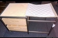 Modern wooden table with two drawers and metal shelf with wire mesh basket under, caster wheels visible. Quilted white mat on top on one side.