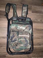 Photo of folding stool with camouflage fabric, black tubular frame, backpack straps, and front storage pockets including zippered compartments.