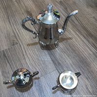 Top down view of three-piece silver-plated coffee set on wood floor. Includes coffee pot, creamer, and sugar bowl with handles and lids.