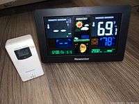 Newentor black digital weather station showing color display with remote sensor temperature, weather forecast icons, indoor temperature, atomic clock, and other data. Included white wireless remote sensor beside it.