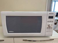 Front view of white Panasonic Genius microwave showing closed door, control panel with digital display, buttons and knob.