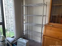 Full view of five-tier metal wire shelving unit placed next to wooden cabinet and window overlooking urban buildings. Unit has chrome finish and is empty.