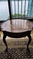 Front view of the wooden side table showing scalloped top surface with scratches and reflection from window, three cabriole legs visible.