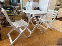 Three-piece white painted wood folding patio furniture set in a room showing table and two chairs with some visible paint wear.