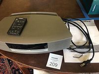 Photo showing Bose Wave Music System unit on a wooden table with power cord and a user booklet alongside a lot label, remote control placed on top of the unit.