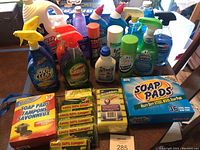 Photo showing many cleaning products arrayed on table including various spray bottles, OxiClean detergent, Comet cleaner, and several packages of scrub sponges and soap pads.