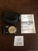 Photo showing Panasonic Lumix DMC-ZS19 black digital camera, protective case, and instruction manual laid out on a surface.