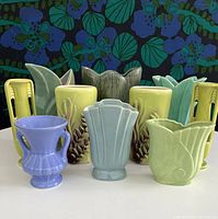 Full group photo showing 10 pottery vases in front of floral fabric backdrop. Vases vary in shapes, colors (blues, greens, yellows), and sizes. Organized in 3 rows.