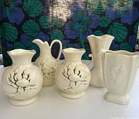 Five vintage cream glazed Red Wing pottery vases arranged in a row, showing magnolia floral decorations on three and varying shapes including a handled ewer and a square vase with embossed detail.