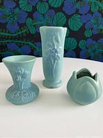 Front view of three turquoise blue Van Briggle pottery pieces against floral background, showing their size and shape.