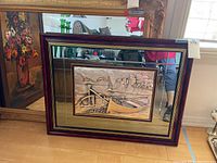 Frontal view of the dimensional metal artwork with a surrounding mirror frame, showing the boat and buildings scene in relief, placed on floor with background items.