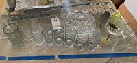 Clear assorted glassware including various drinking glasses, mugs, and vases on a table with tablecloth. Pieces are transparent and colorless, some marked Arcoroc France.
