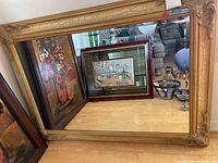 Photo showing large rectangular mirror with ornate gold frame leaning against a wall with paintings reflected in the glass.