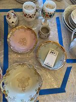 Top-down view of Limoges sugar bowl with lid, three decorated porcelain bowls with gold rims in pink, green and cream, and small milk pot with gold on a lace tablecloth.