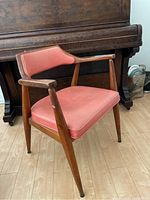 Side angled view of the vintage wood and vinyl Mid-Century Modern style chair placed on a wooden floor with a dark piano in background.
