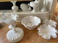 Overview of milk glass pieces on wooden floor and ledge: multiple vases, bowls, candy jar, and swan figurine