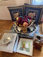 Photo showing two elephant-themed decorative pillows in blue and gold, a wire fruit basket, silver serving trays with artificial fruit, a ceramic vase, and a ceramic serving dish with a colorful vintage design.