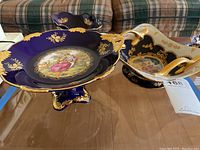 Photo showing the three decorative bowls on a glass table. Visible is the large cobalt blue pedestal bowl with gold leaf accents, the square bowl with gold handles, and a smaller bowl in the background.