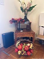 Wooden side table with lamp and decorative items on top, paper shredder to the left, basket of faux fruits in front