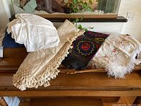 Five tablecloths and linens arranged on a wooden surface showing various fabric colors and textures including white, cream, and colorful embroidered fabric.