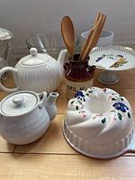 View of two ceramic teapots, white bundt cake plate with floral pattern, glass bowl, wooden utensils in brown ceramic holder, decorative bird plate.