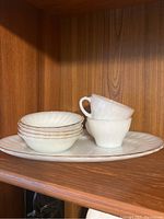 White milk glass pieces including stacked teacups, small bowls, and large platter shown on wooden shelf