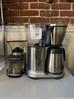 Front view of Breville coffee maker with digital display and Cuisinart coffee grinder side by side on a white surface against a brick wall.