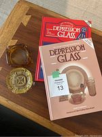 Two hardcover copies of The Collector's Encyclopedia of Depression Glass and two vintage glass ashtrays on a wooden table.