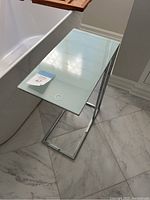 View of the glass top and silver metal frame side table positioned on a tiled floor next to a bathtub.