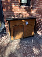 Front view of the cabin-style dog house with wooden slat panels and metal frame on a brick patio.