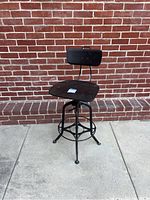 Front view of the industrial style counter stool showing the wooden seat, backrest, and metal frame on concrete floor with brick wall background